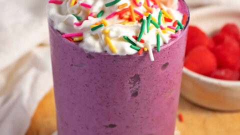 Homemade Grimace Shake Recipe with Whipped Cream and Sprinkles Featuring Raspberries in the Background