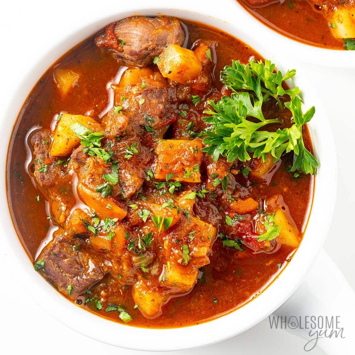 Beef stew on a white bowl with tomato sauce and potatoes garnished with fresh parsley. 