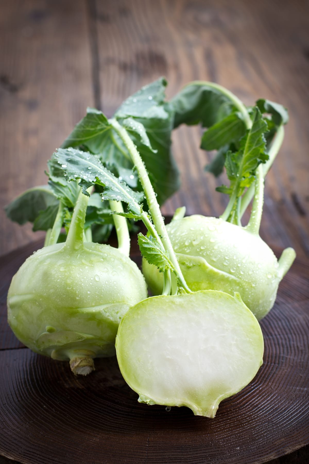 What Is Kohlrabi? (All You Need to Know) featuring Two Whole and One Sliced Dewy Fresh Kohlrabi on a Wooden Table