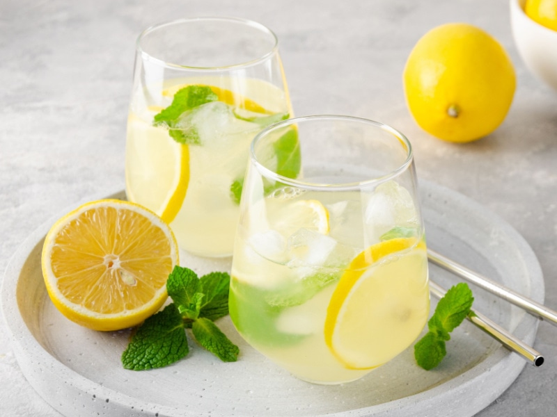 Two Traditional Lemonades with Lemon Slices, Mint, and Ice in a Glass on a Stone Serving Tray with Metal Straws and More Mint and Lemon 