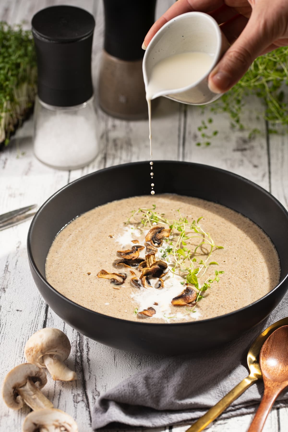 How to Thicken Soup (8 Easy Methods!) featuring Milk Poured into a Bowl of Mushroom Soup with Sauteed Mushrooms and Microgreens on a Wooden Table with More Mushrooms and a Gold Spoon Around the Bowl