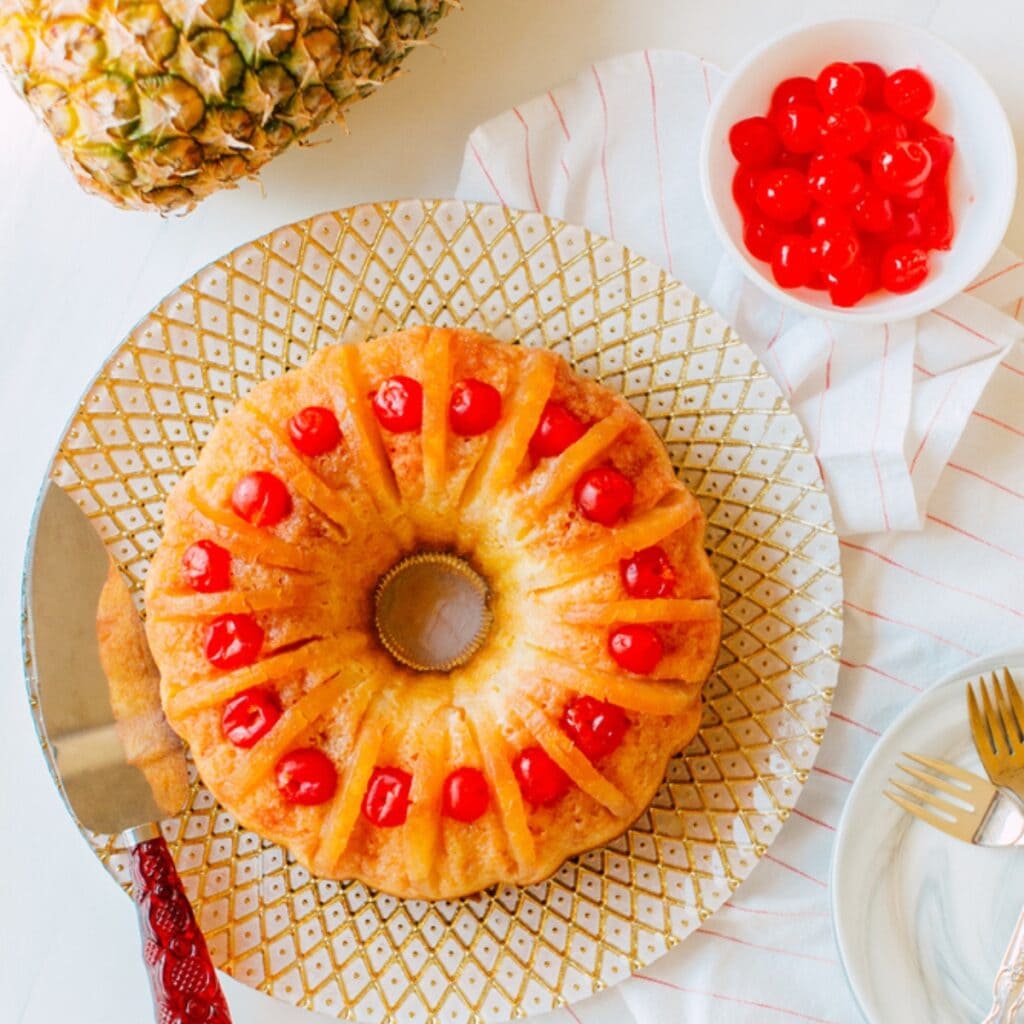 Top View of Whole Pineapple Upside-Down Bundt Cake Served on an Elegant White and Gold-Patterned Serving Plate with Serving Spatula and Cherries and Forks to the Side