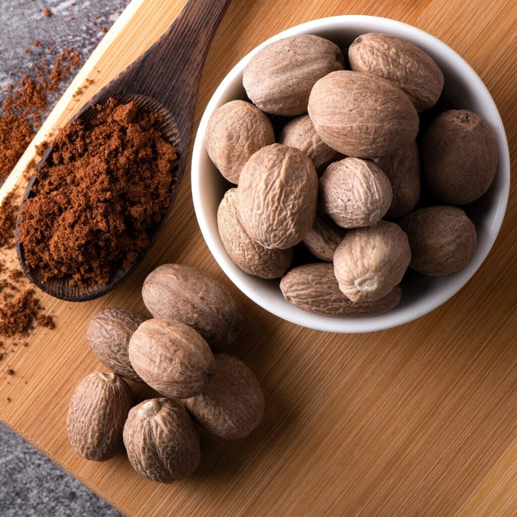 What Is Nutmeg? (Its Flavor, Uses, & More!) featuring Whole Raw Organic Nutmeg and Nutmeg Powder on a Wooden Board