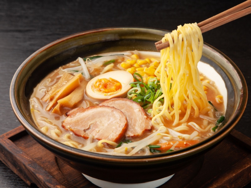 Sapporo Ramen in a Bowl with Pork, Egg, Bamboo Shoots, Corn, Bean Sprouts, and Scallions with Noodles Held By Chopsticks