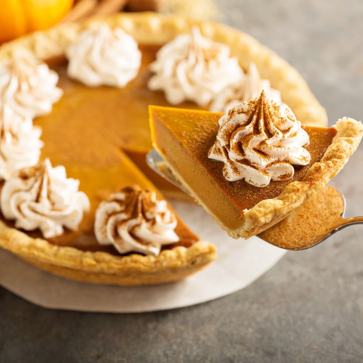 Slice of Pumpkin Pie with Whipped Cream and Cinnamon on a Pie Spatula in the Air, with the Rest of the Pumpkin Pie on a Board on the Table