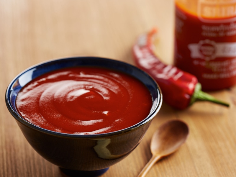 Bowl of Sriracha, Fresh Chili, Bottle of Sriracha Sauce, and Wooden Spoon on a Wooden Table