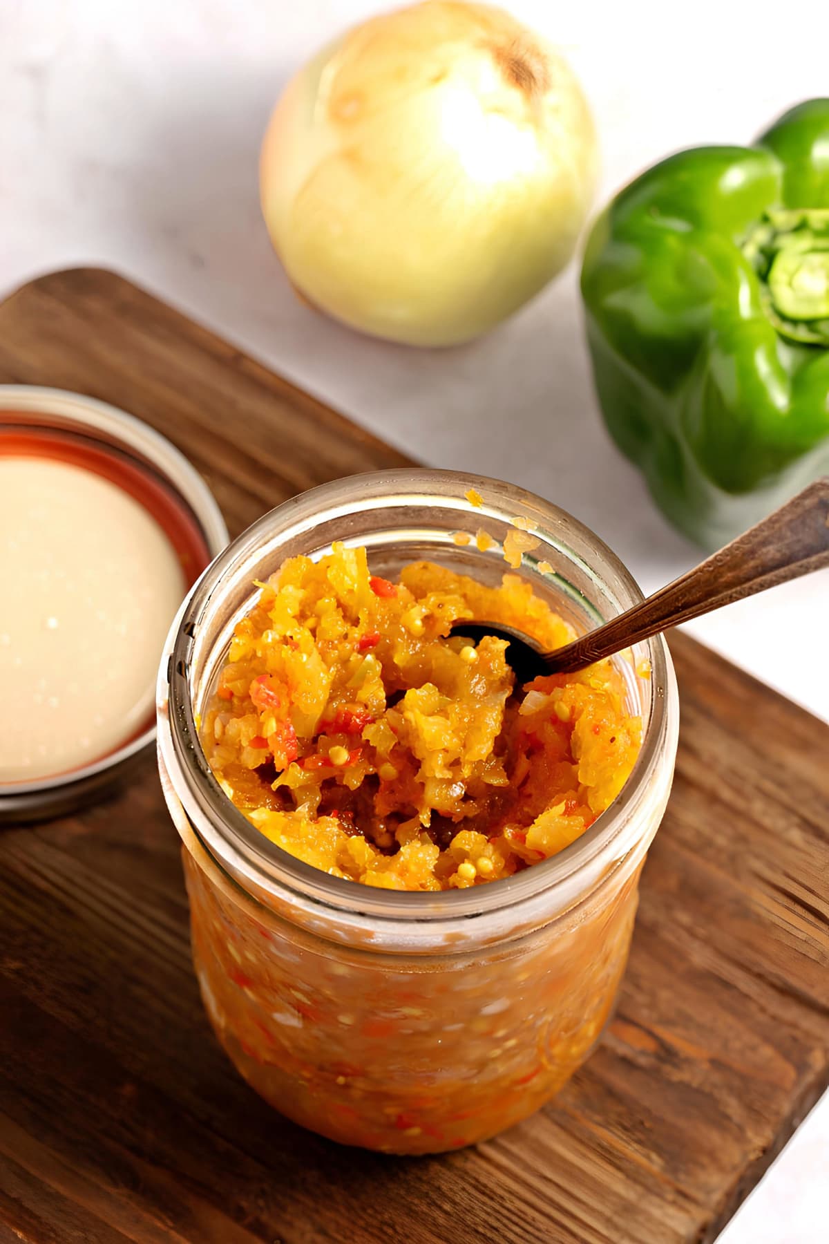 The Best Green Tomato Relish (Easy Recipe) featuring Sweet and Tangy Green Tomato Relish in a Mason Jar with a Spoon and Pepper and Onion in the Background