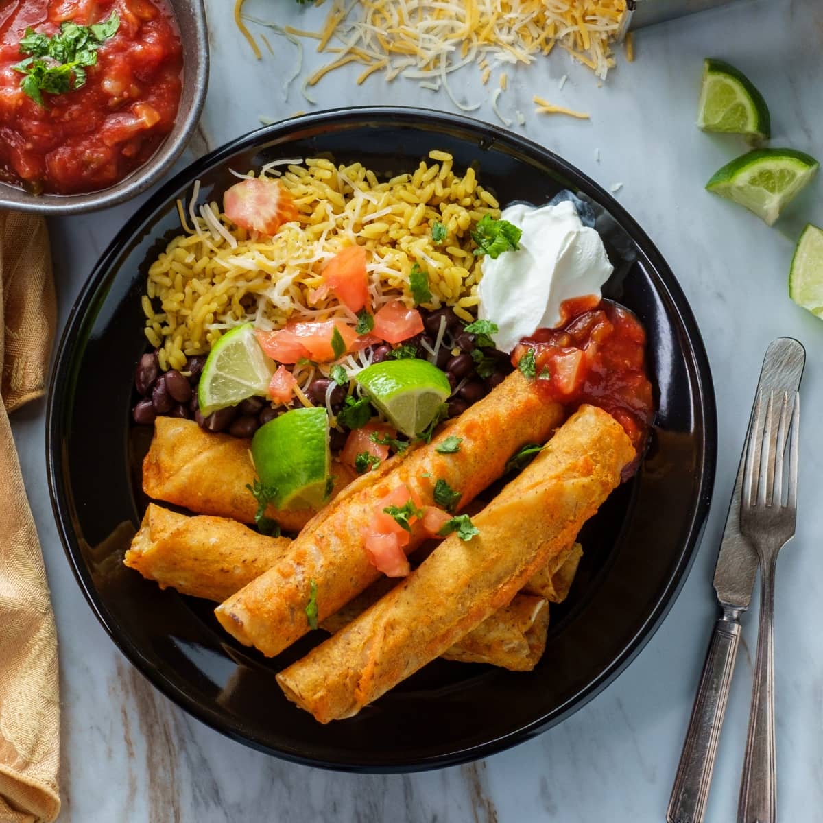 Taquitos Served With Rice, Cheese, Beans, Limes, Sour Cream, and Salsa on a Plate with Knife and Fork Beside It