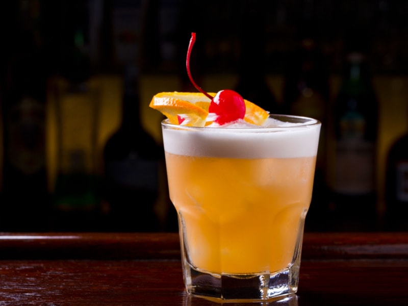 A Glass of Tequila Sour Garnished with Cherry and an Orange Slice on a Wooden Table in a Dark Room