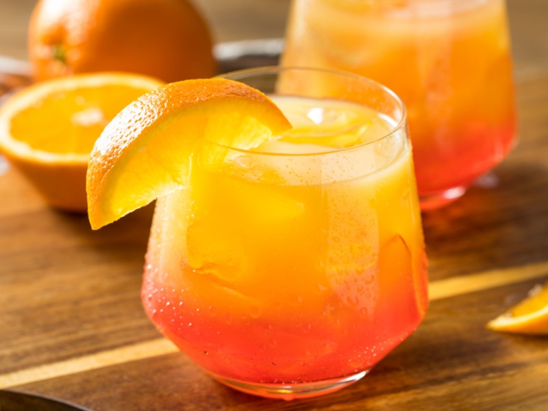 2 Glasses of Boozy Refreshing Tequila Sunrise Cocktail with Grenadine and Ice Garnished with Orange Slices o Wooden Table with Sliced Oranges 