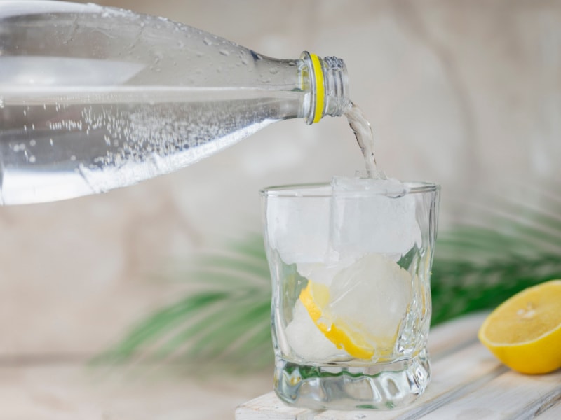 A Bottle of Tonic Water Pouring into a Glass with Ice and Lemon Slice and More Lemon in the Background and Some Blurry Greenery
