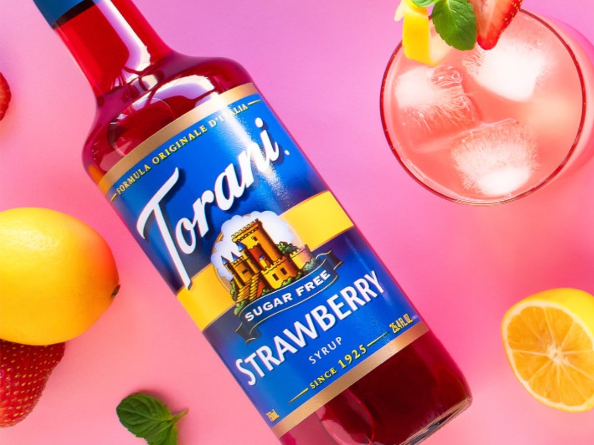 Bottle of Torani Sugar-Free Strawberry Syrup and Ice Cold Strawberry Drinks with Strawberries and Lemons in the Background