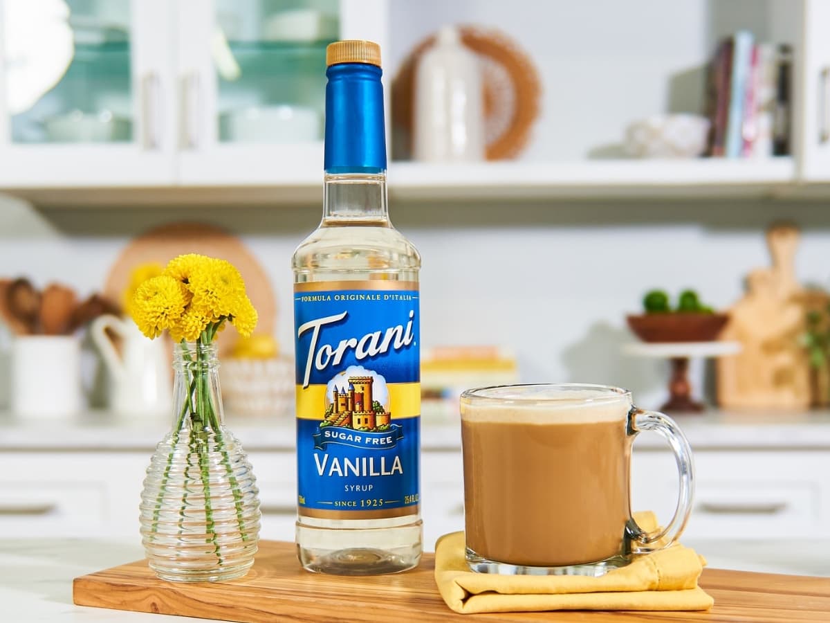 Bottle of Torani Sugar-Free Vanilla Syrup on Wooden Cutting Board with a Glass Mug of Coffee to the Right and a Vase of Yellow Mini Chrysanthemums to the Left