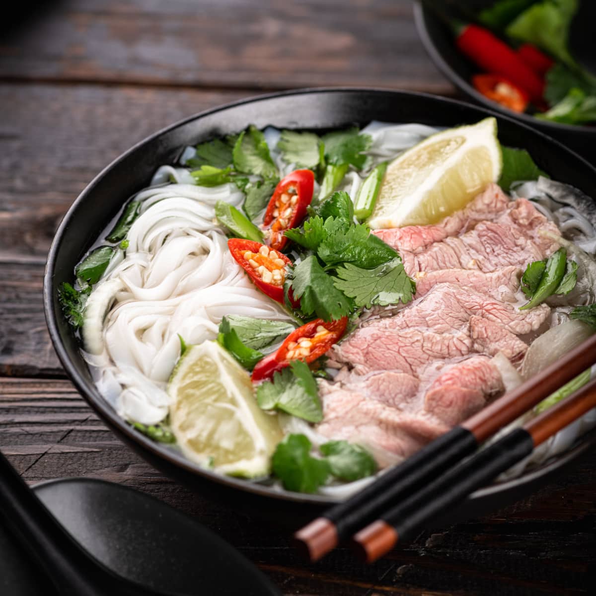 Pho vs. Ramen- The Main Differences featuring Vietnamese Pho Noodle Soup with Rice Noodles, Beef, Cilantro, Chiles, and Lemon in a Bowl with Chopsticks