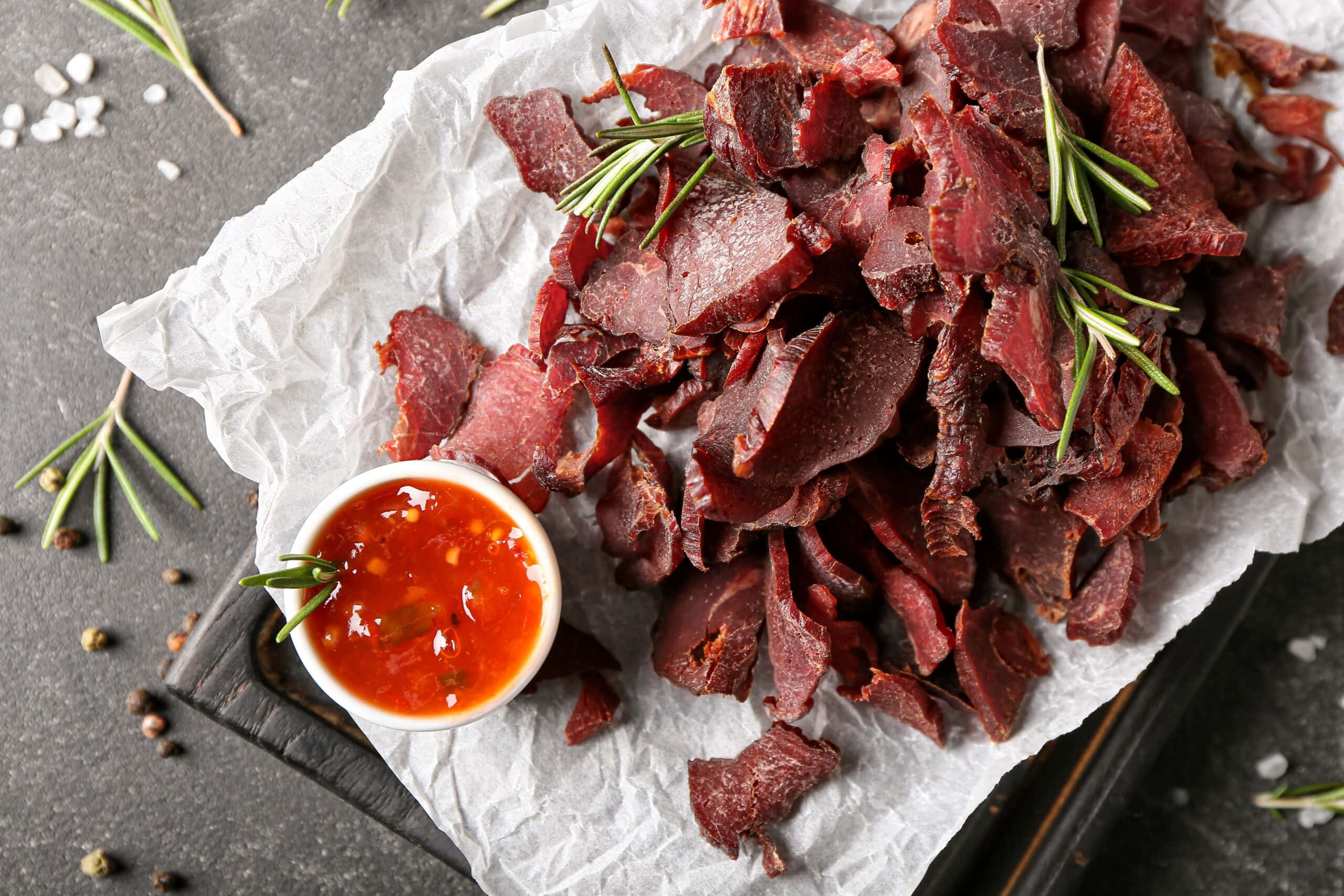 https://insanelygoodrecipes.com/wp-content/uploads/2023/09/Beef-Jerky-with-Sweet-and-Sour-Sauce-Garnished-with-Rosemary-Leaves-on-a-Wooden-Cutting-Board-with-Parchment-Paper-scaled.jpg