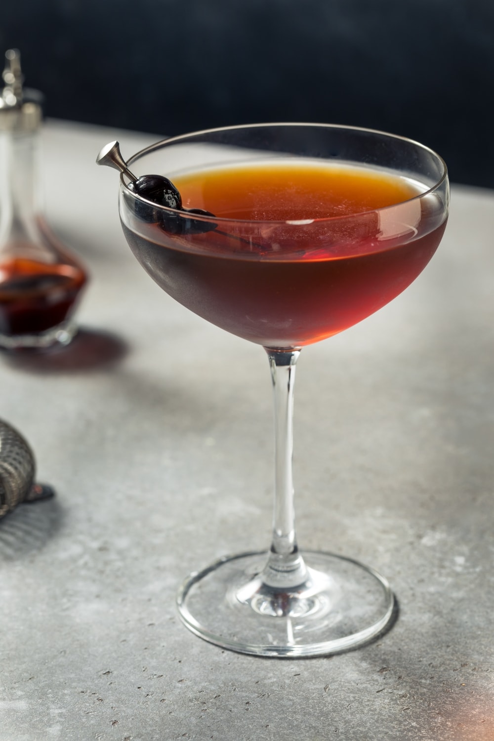 Glass of Refreshing and Boozy Black Manhattan Cocktail with Black Cherry Garnish and Cocktail-Making Tools in Background
