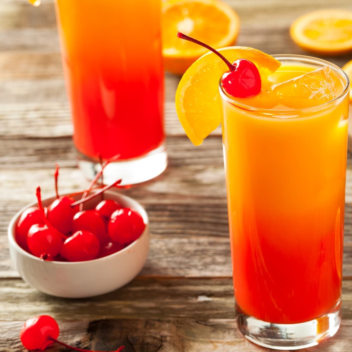 Two Tall Glasses of Refreshing Tequila Sunrise with Cherries and Orange Slices and a Bowl of Maraschino Cherries to the Side
