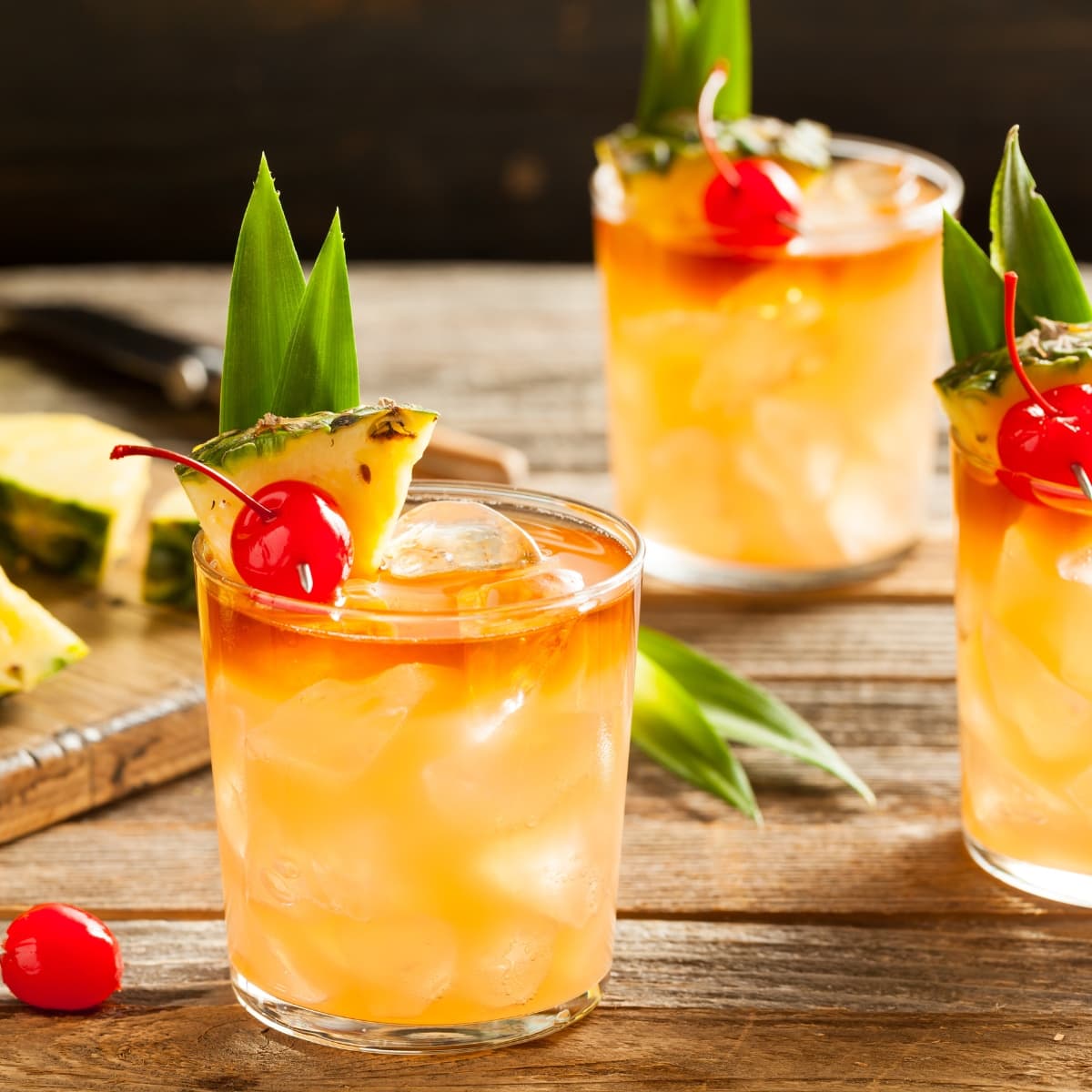 Three Glasses of Mai Tai Cocktail with Cherry and Pineapple Garnish on a Wooden Table with a Wooden Cutting Board in the Background with Cut Pineapple Slices