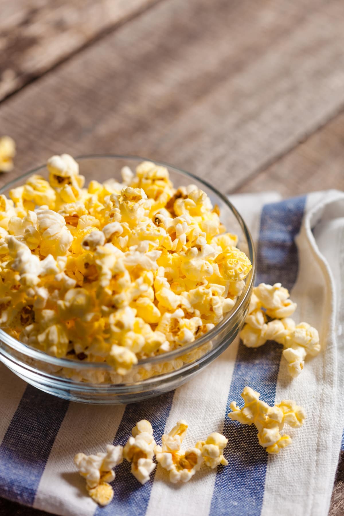 The Best Popcorn Toppings- 15 Ideas To Try featuring Homemade Popcorn in a Glass Bowl on a Wooden Table with a Blue and White Dishcloth