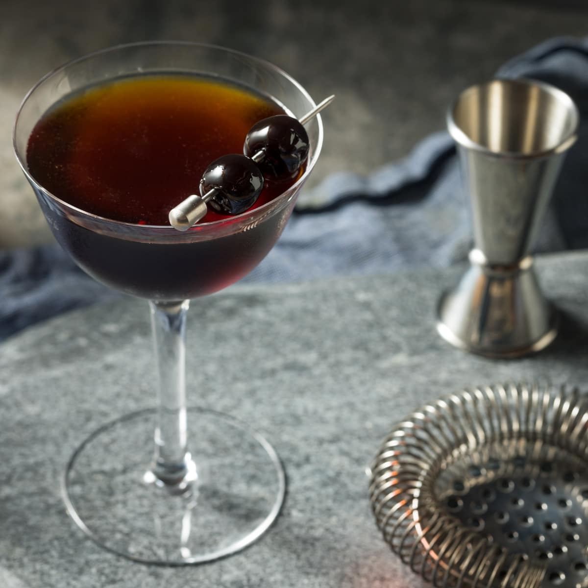 Mixer, Strainer, Shot Measurer, and a Glass of Black Manhattan Cocktail with a Black Cherry Garnish
