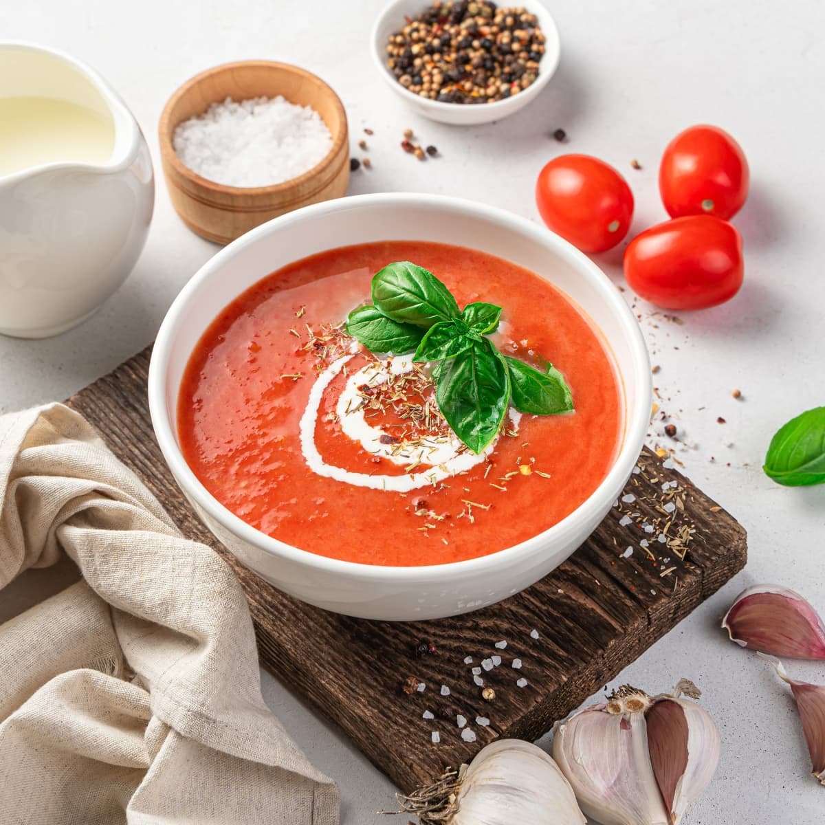 A bowl of tomato sauce garnished with cream and fresh basil leaves surrounded by onion, garlic cloves, salt, tomato and pepper on a table. 