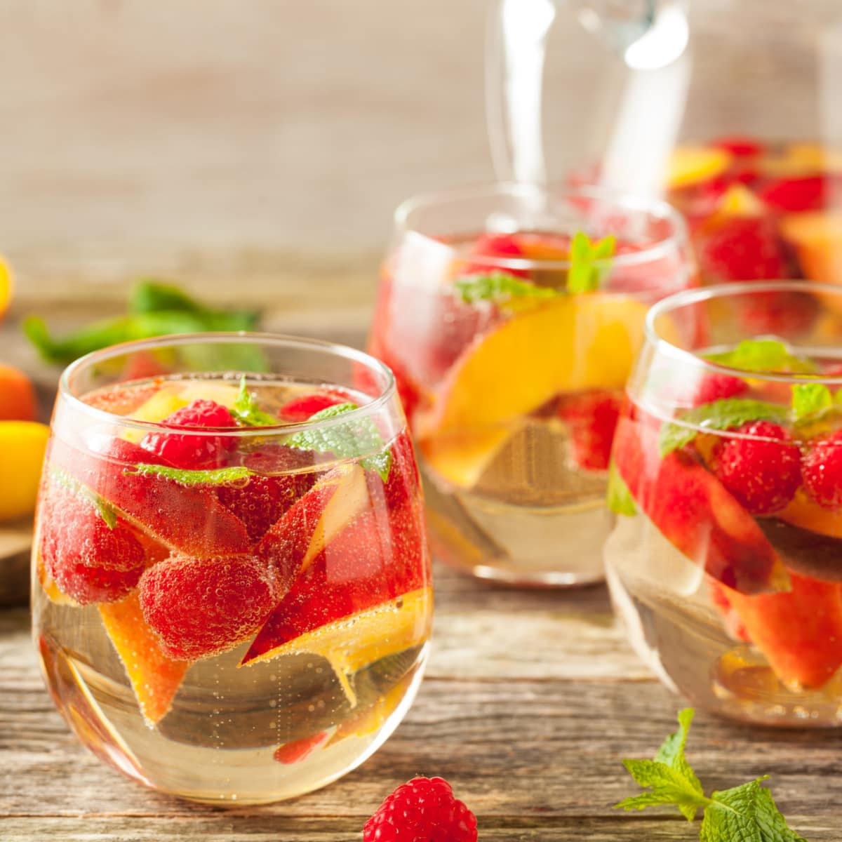 A pitcher and three glasses of white sangria on a wooden table with fresh raspberries and peaches