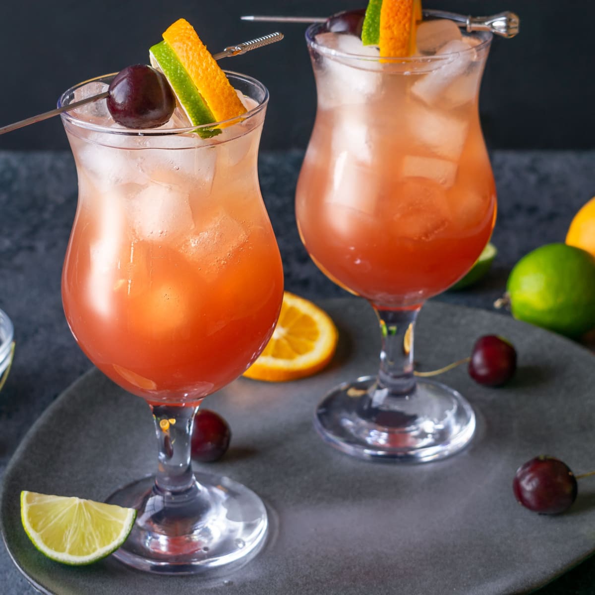 Two Hurricane Cocktails Served in Poco Grande Glasses, Garnished with A Cherry, Lime Wheel, and Orange Wheel on a Gray Platter with Cherries, Limes, and Oranges All Around the Platter