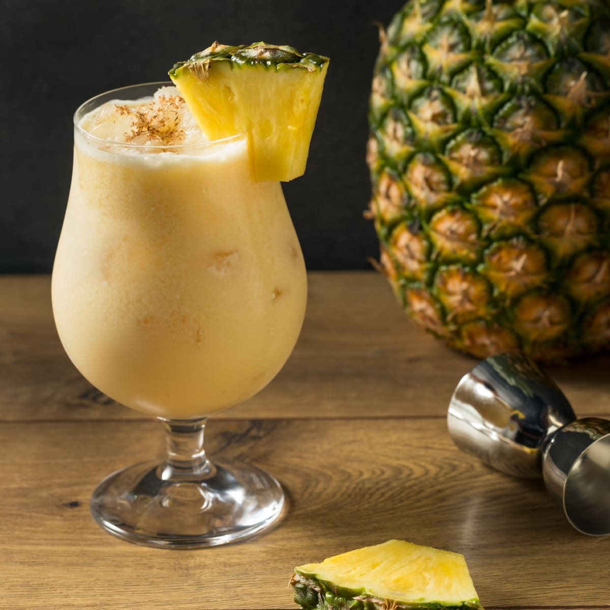 Whole pineapple and a glass of Painkiller Cocktail garnished with pineapple slice on a wooden table with a jigger and a whole pineapple to the right side