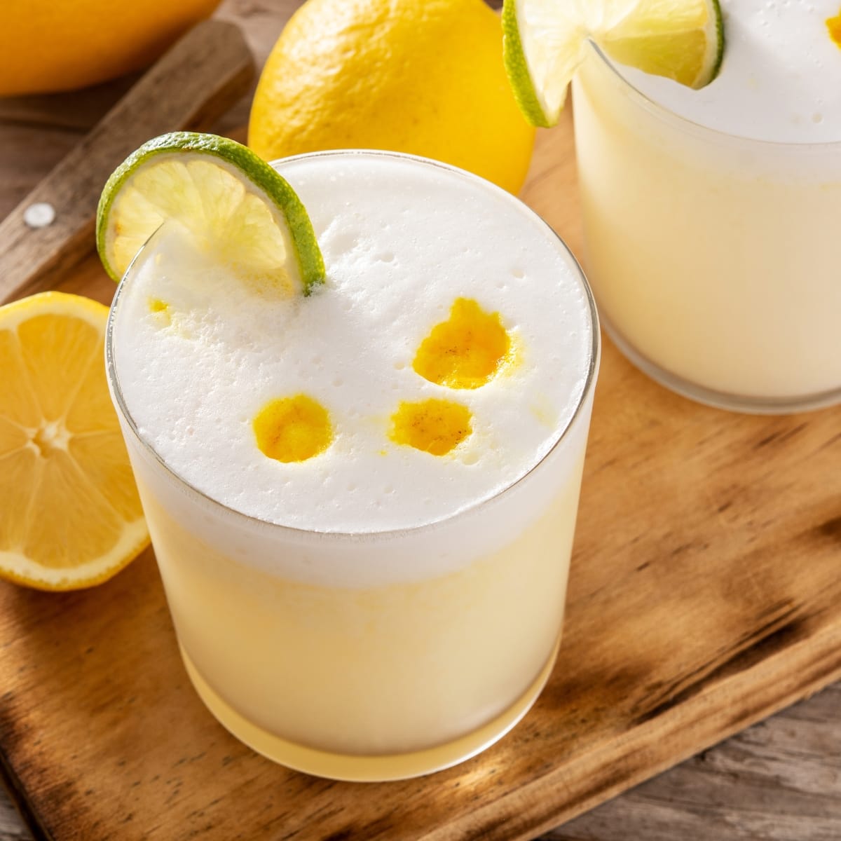 Two Glasses of Pisco Sour Garnished With Slice of Lime Wheel on a Cutting Board with Sliced Lemons
