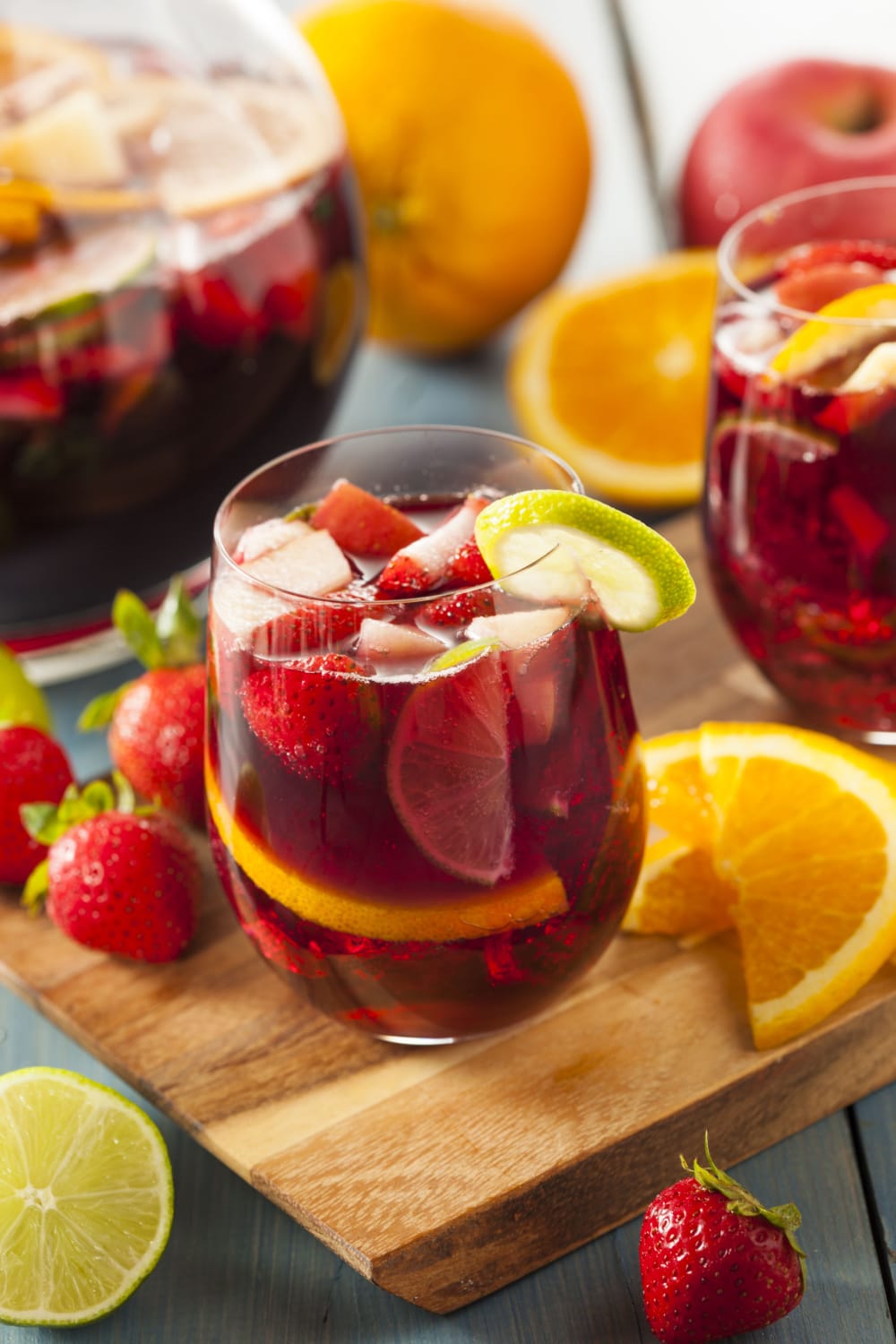 Red sangria cocktail served in glasses with fresh apples, oranges, strawberries and lime on top of wooden cutting board. 