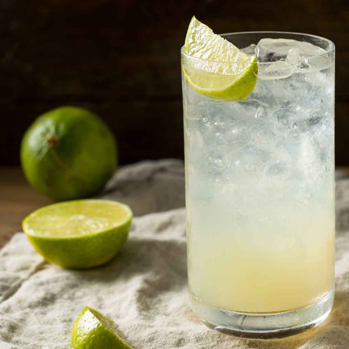 Refreshing glass of ice cold Gin Rickey garnished with sliced green lime, and whole and sliced limes on side