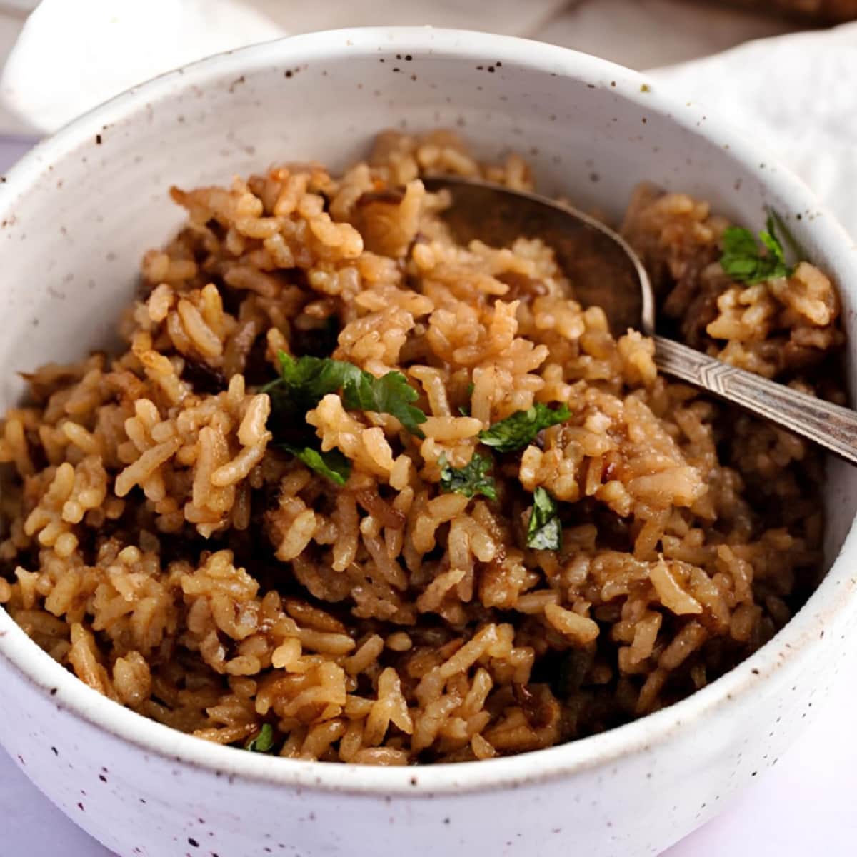 Angled view of brown colored stick of butter rice, with a spoon, topped with few parsley leaves