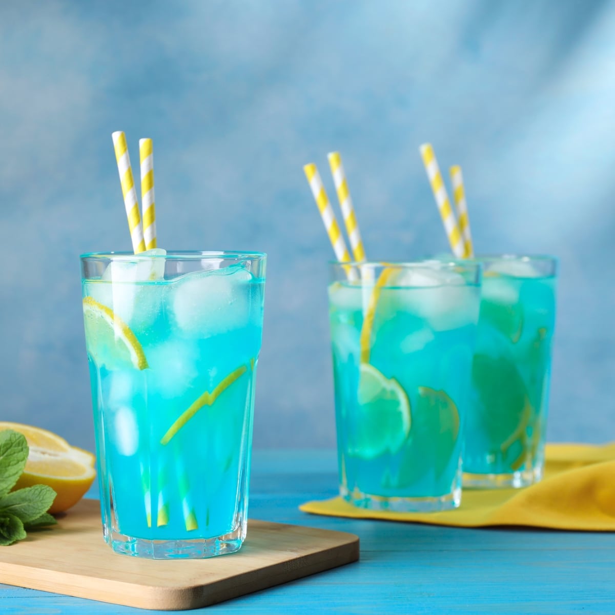 Three Glasses of ice-filled Blue Lagoon Cocktails with yellow-striped straws and  lemon slices