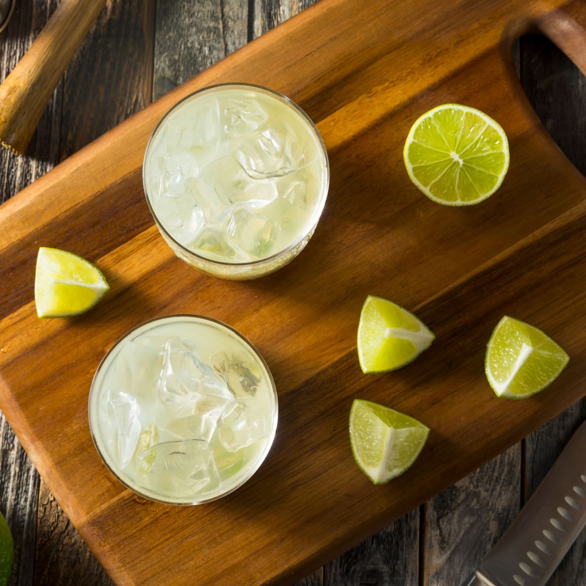 Top view of two glasses of Caipirinha Cocktail and slices of lime on a wooden board with a knife to the side