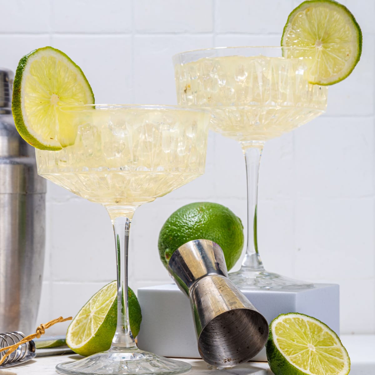 Two glasses of gimlet cocktail garnished with lime wheel placed neatly on a countertop with a jigger, shaker lime slices