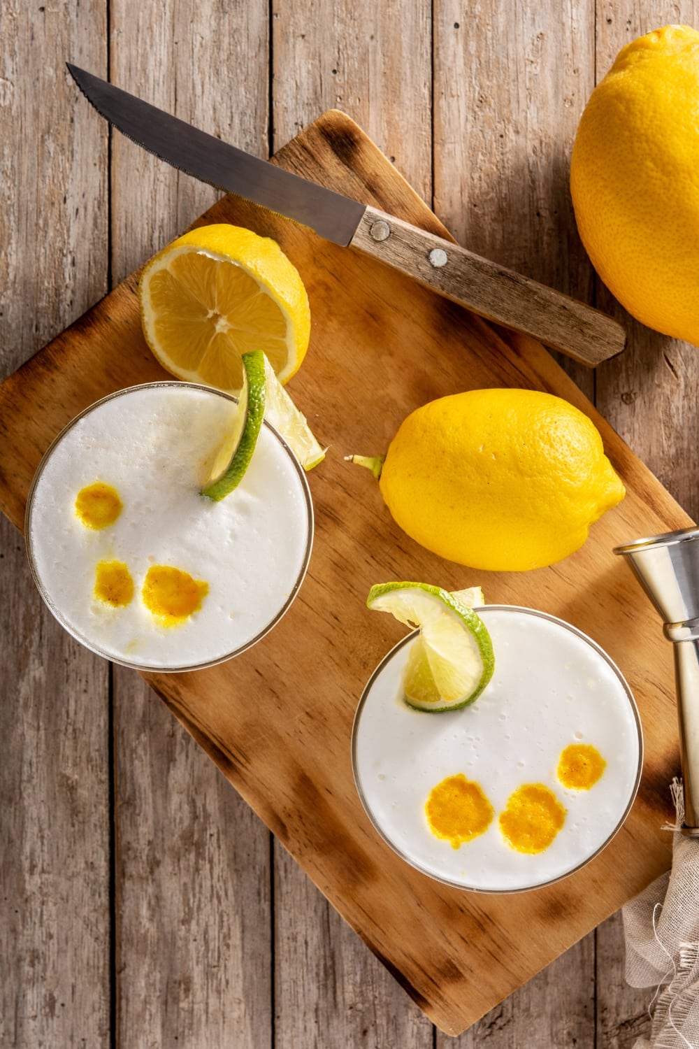 Two Glasses of Foamy Pisco Sour Top View on a Wooden Cutting Board with Lemons, a Knife, and a Shot Measurer
