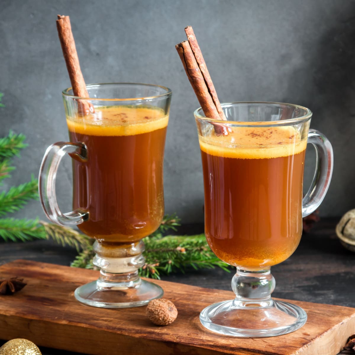 Two Mug Glasses of Hot Buttered Rum with a cinnamon Stick Garnish on a Wooden Board
