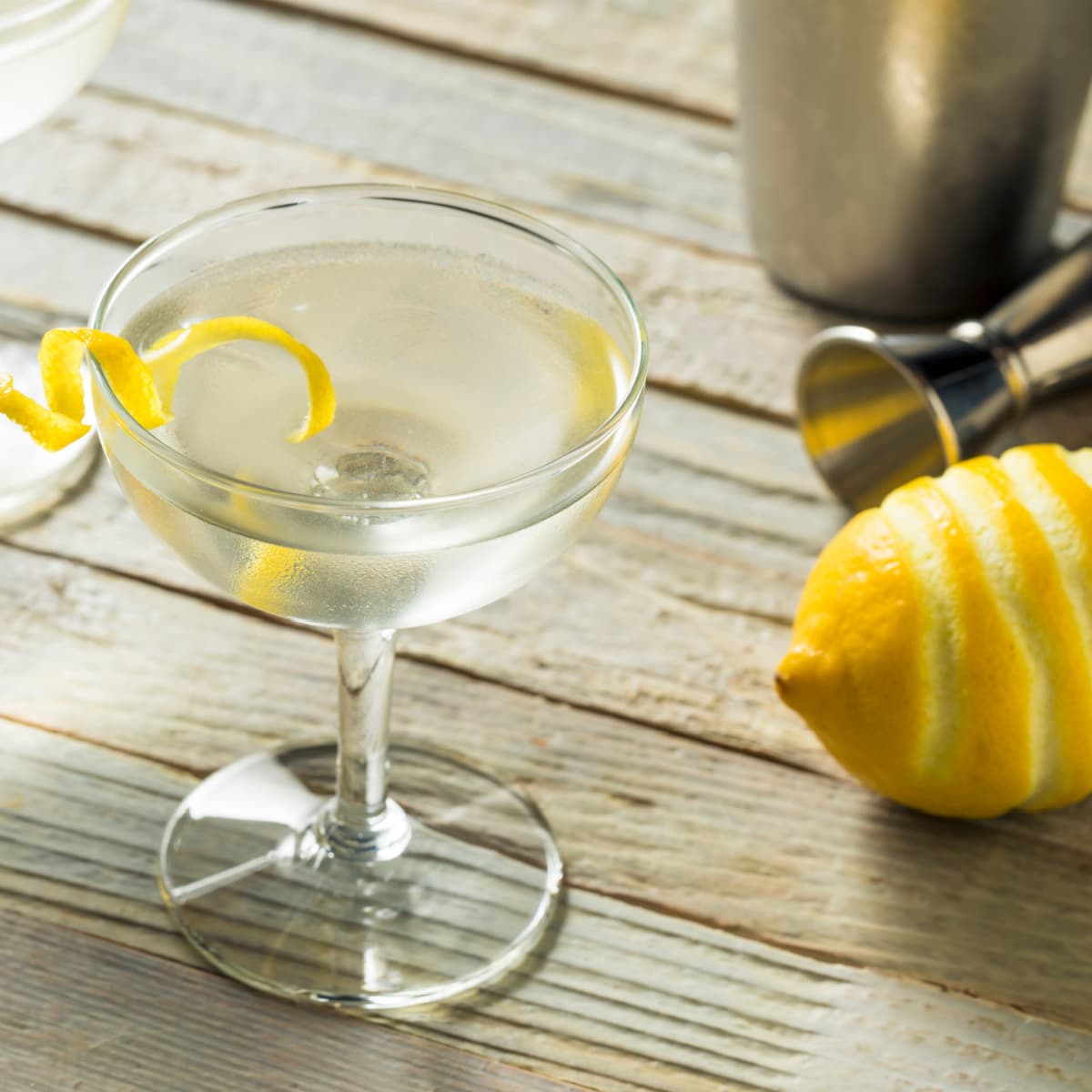 A glass of Vesper Martini garnished with peeled lemon, with jigger and shaker on a wooden table
