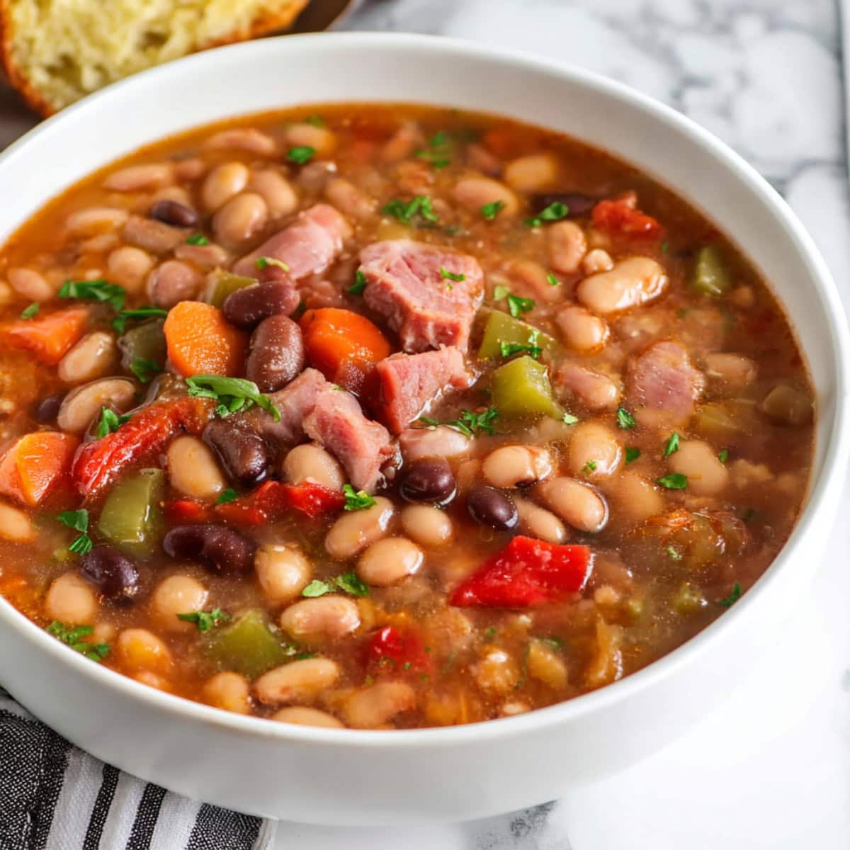 A close-up of 10-bean soup with red and green bell peppers, ham hock and garnished with herbs.