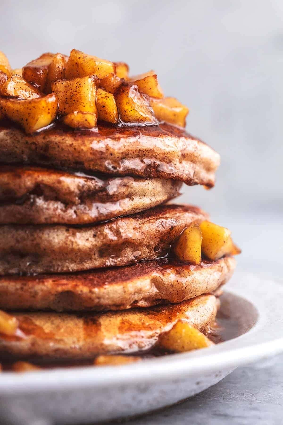 Stack of apple cinnamon pancakes drizzled with maple syrup and sprinkled with cinnamon powder topped with apple slices. 