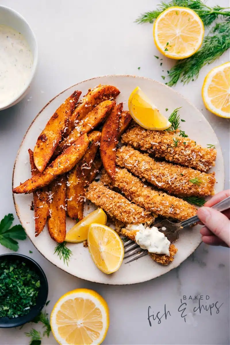 Baked breaded fish sticks with French fries served on plate with lemon wedge. 