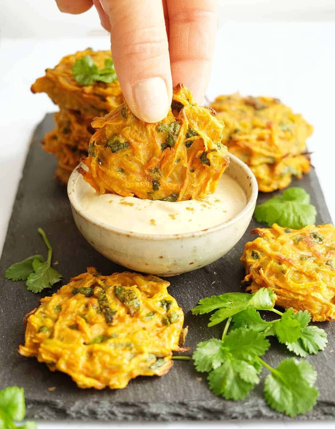 Carrot Fritters dipped on a bowl of hummus dip with fresh cilantro on black stone tablet