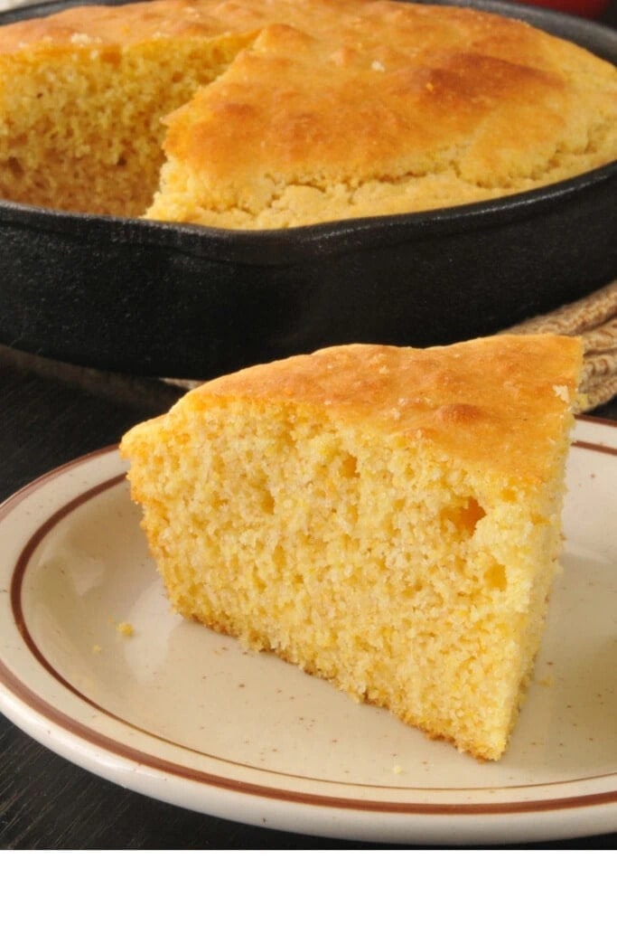 A slice of cornbread on a plate with freshly bake cornbread on a cast iron pan in the background