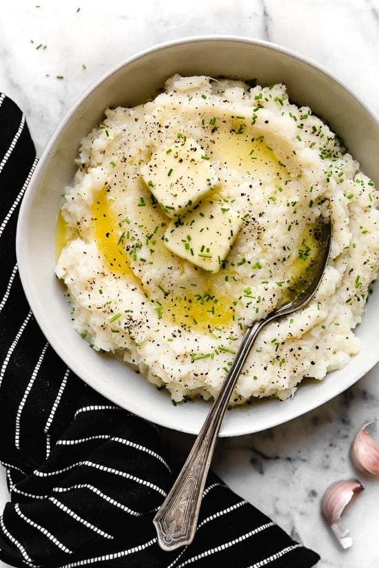 Mashed cauliflower puree in a bowl garnished with rosemary and melted butter with spoon. 