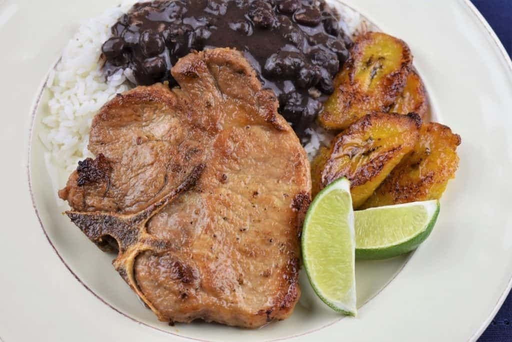 Cuban pork chops served with rice garnished with lime wedge, potatoes and black beans. 