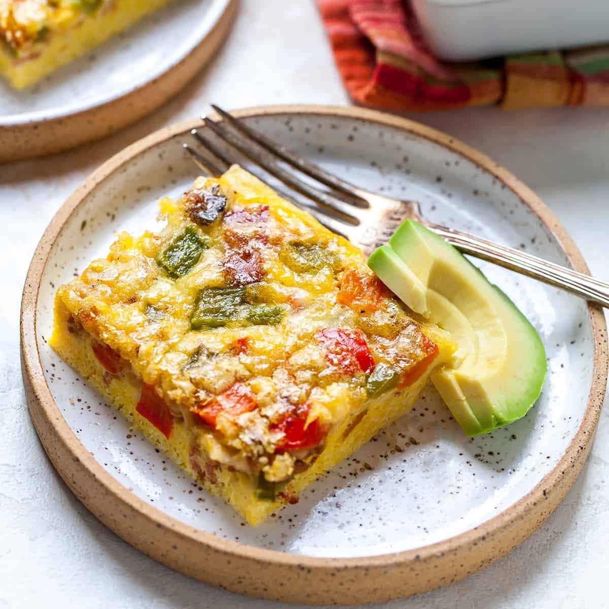 Slice of egg casserole with bacon, eggs, vegetables and cheese on a saucer with slices of avocado