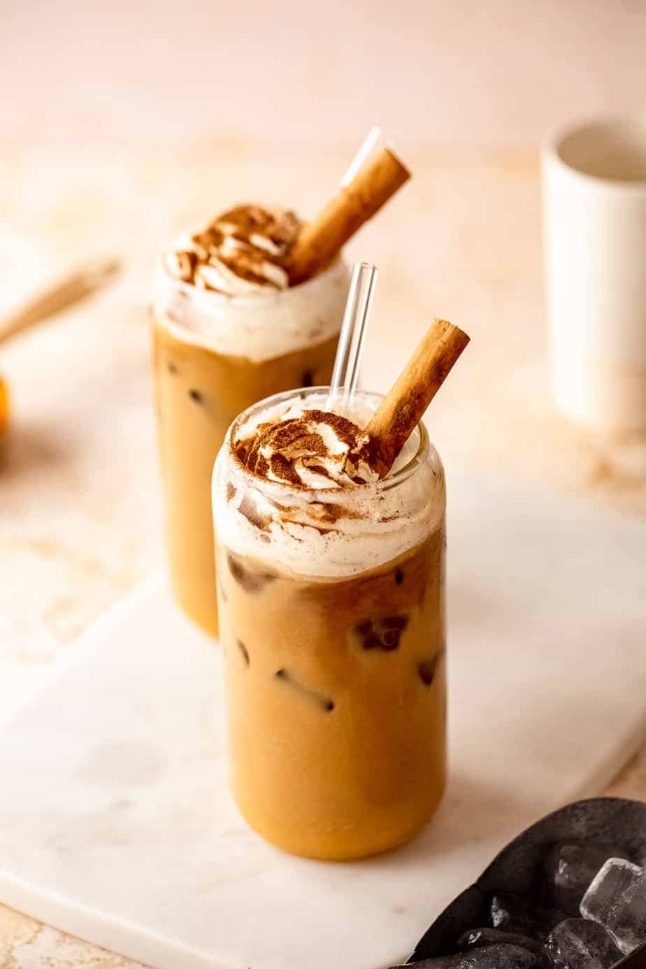 Two glasses of iced pumpkin chai latte garnished with cinnamon stick and powder. 