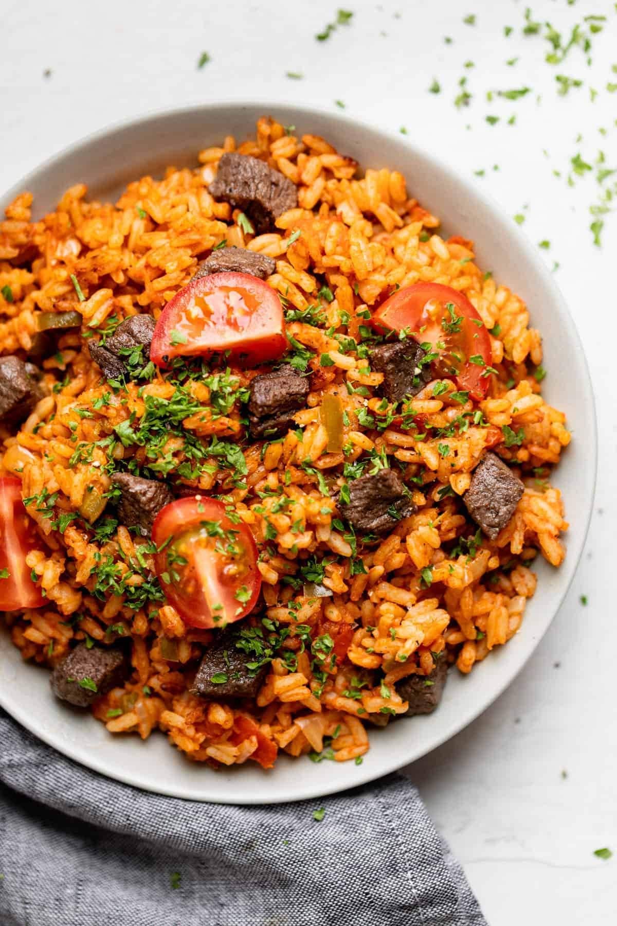Jollof rice in a white bowl with beef, tomato slices and chopped herbs. 