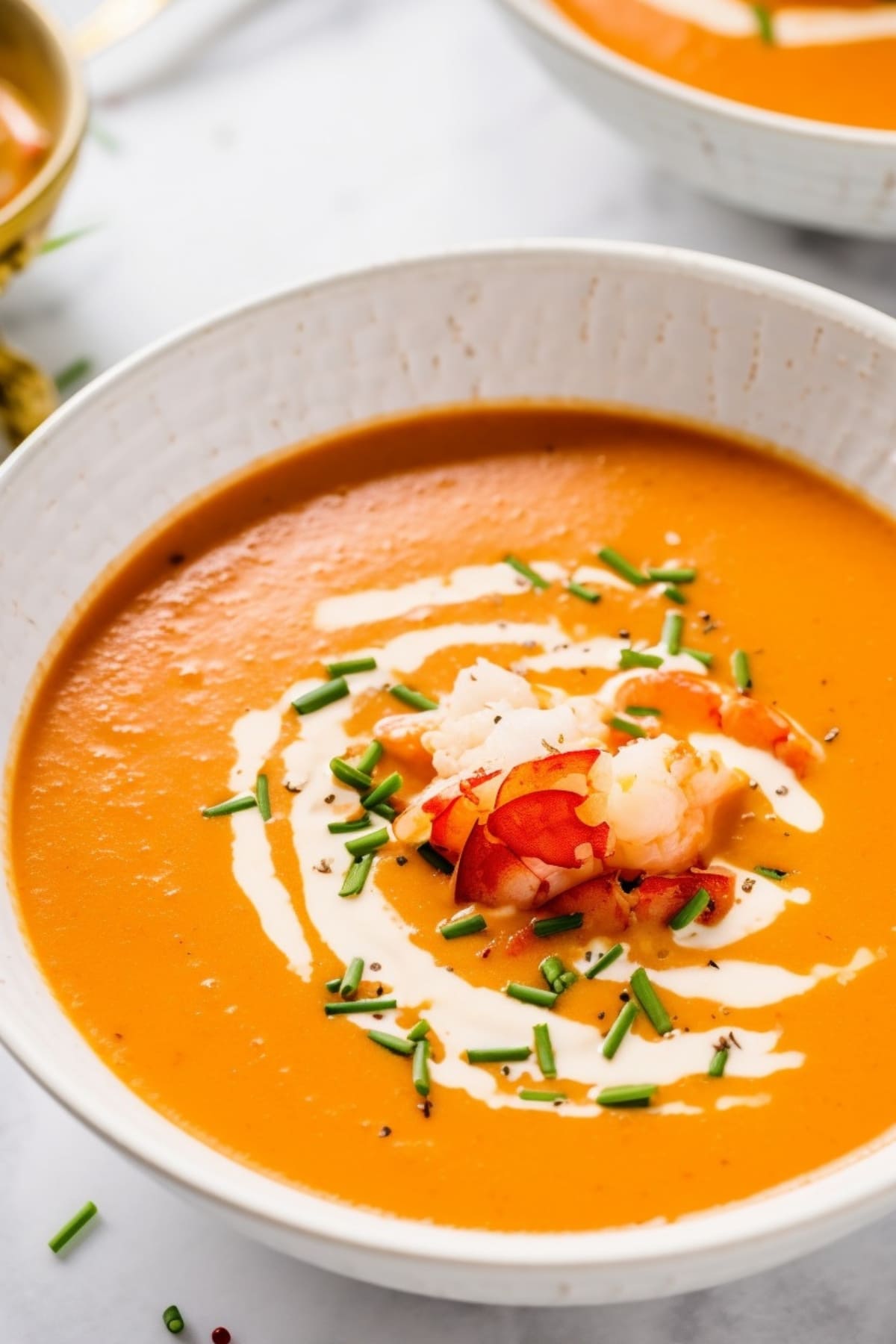 Creamy lobster bisque served in a white bowl drizzled with cream garnished with lobster meat and chopped leeks. 