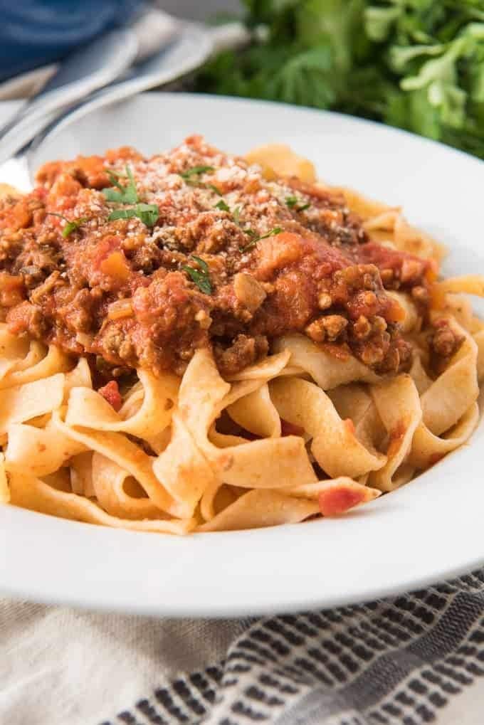 Pasta topped with Bolognese sauce, garnished with cheese and chopped parsley served on a white plate. 